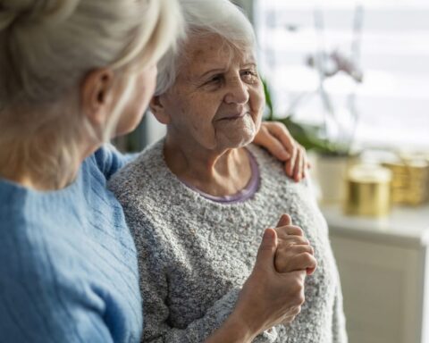 Zwei ältere Frauen sitzen zusammen in einer hellen Wohnung und halten sich an den Händen.