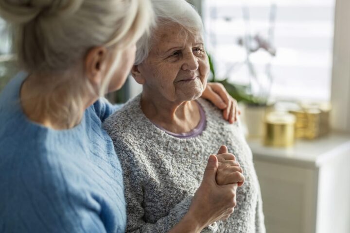 Zwei ältere Frauen sitzen zusammen in einer hellen Wohnung und halten sich an den Händen.