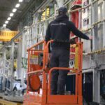Ein Arbeiter steht auf einer orangefarbenen Hubarbeitsbühne in einer Fabrikhalle neben einer großen Industrieanlage.