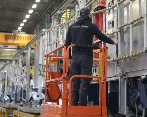 Ein Arbeiter steht auf einer orangefarbenen Hubarbeitsbühne in einer Fabrikhalle neben einer großen Industrieanlage.