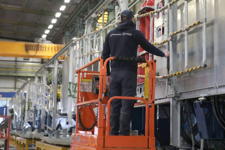 Ein Arbeiter steht auf einer orangefarbenen Hubarbeitsbühne in einer Fabrikhalle neben einer großen Industrieanlage.