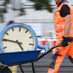 Bauarbeiter in orangefarbener Schutzkleidung schiebt eine Schubkarre mit einer großen Uhr.