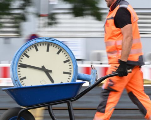 Bauarbeiter in orangefarbener Schutzkleidung schiebt eine Schubkarre mit einer großen Uhr.