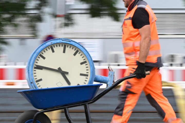 Bauarbeiter in orangefarbener Schutzkleidung schiebt eine Schubkarre mit einer großen Uhr.