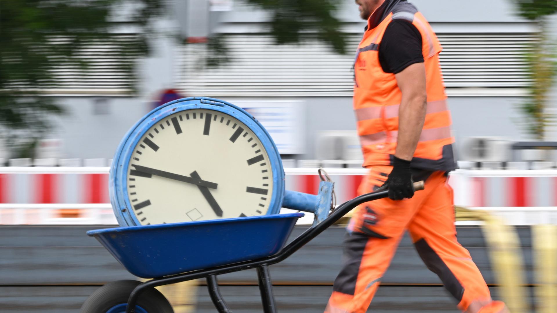 SPD-Gruppen ziehen rote Linien bei Arbeitszeitreform der Koalition 1 Bauarbeiter in orangefarbener Schutzkleidung schiebt eine Schubkarre mit einer großen Uhr.