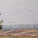 Blick über eine staubige Landschaft mit Bäumen im Vordergrund und einer fernen, verschwommenen Bebauung unter grauem Himmel.