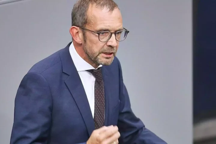 Mann im Anzug und mit Brille spricht vor neutralem Hintergrund.