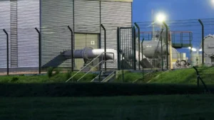 Blick auf eine Industrieanlage mit Rohrleitungen hinter einem Zaun.