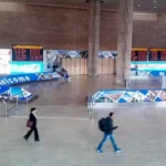 Innenhalle eines Flughafens mit Anzeigetafeln, Begrüßungsbeschilderung und wenigen Passanten.