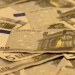 Fünf-Euro-Scheine liegen gestapelt und verstreut auf einer Fläche.