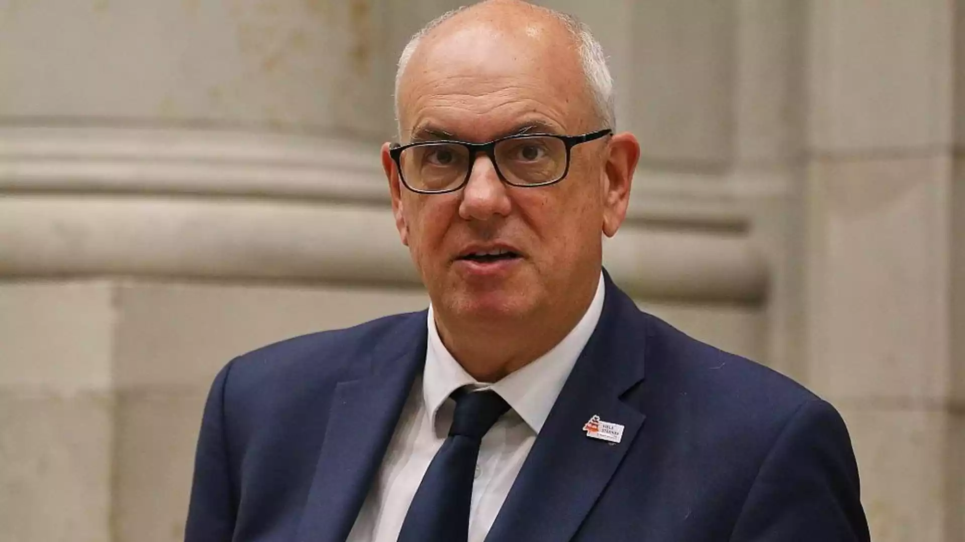 Ein älterer Mann mit Brille und dunklem Anzug spricht vor hellem Hintergrund.