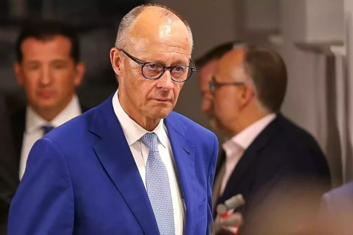 Ein Mann im blauen Anzug und mit Brille steht in einem Innenraum, im Hintergrund sind weitere Personen unscharf zu sehen.