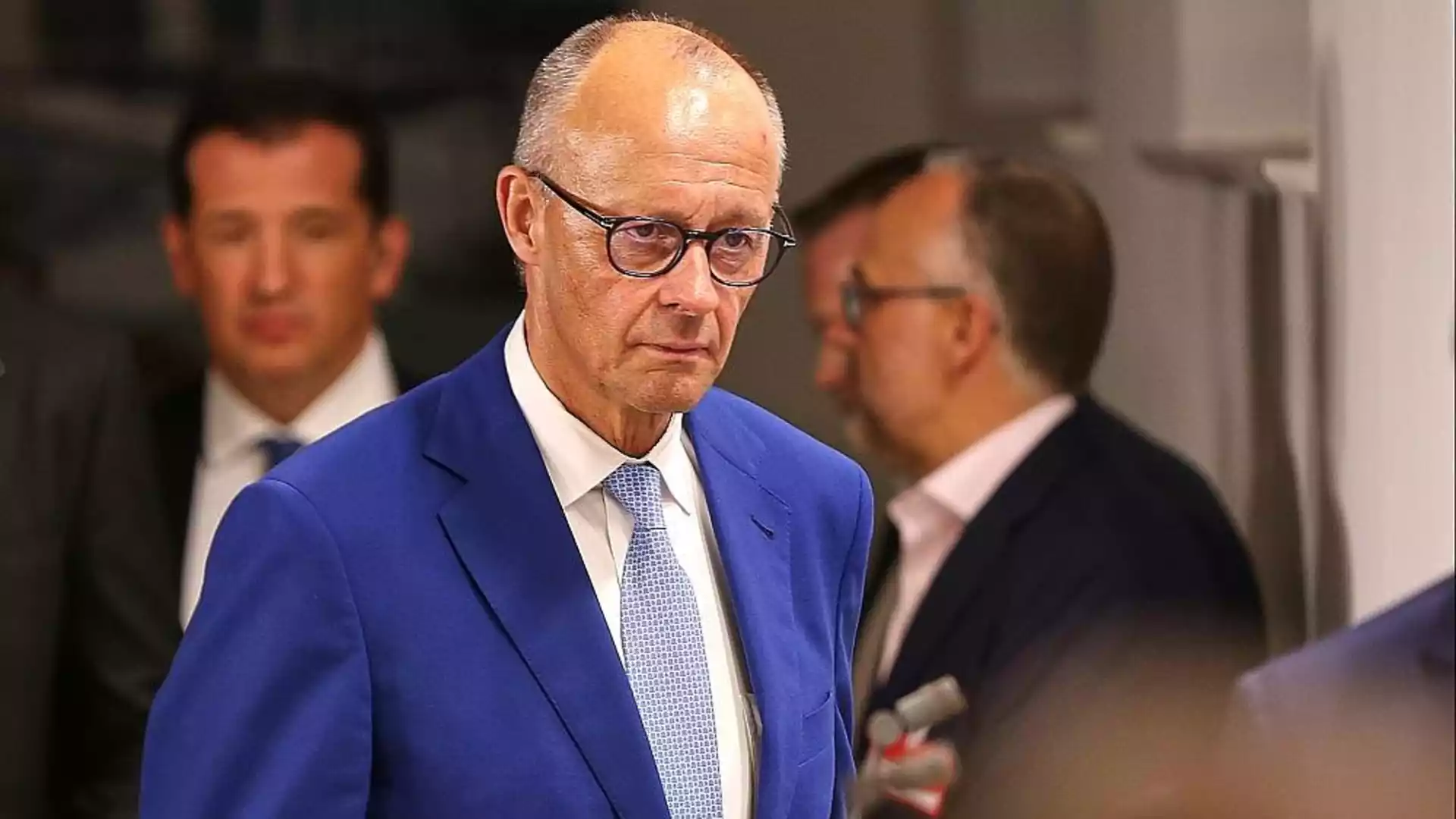 Ein Mann im blauen Anzug und mit Brille steht in einem Innenraum, im Hintergrund sind weitere Personen unscharf zu sehen.