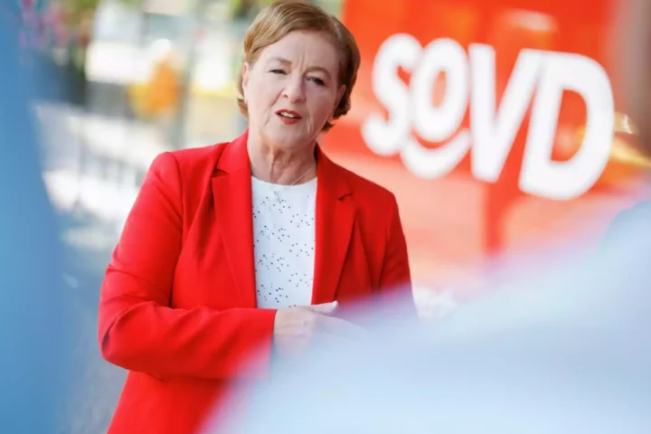 Eine Frau in rotem Blazer spricht vor einem orangefarbenen Hintergrund mit dem Schriftzug „SoVD“.