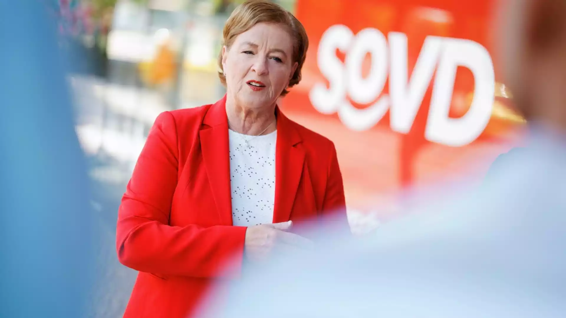 SoVD fordert Erhalt der Verhinderungspflege für pflegende Angehörige als Entlastung 1 Eine Frau in rotem Blazer spricht vor einem orangefarbenen Hintergrund mit dem Schriftzug „SoVD“.