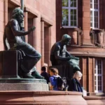 Vor einem roten Sandsteingebäude stehen bronzene Sitzstatuen, davor gehen mehrere Studierende vorbei.