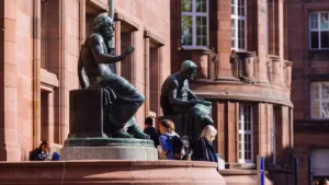 Vor einem roten Sandsteingebäude stehen bronzene Sitzstatuen, davor gehen mehrere Studierende vorbei.