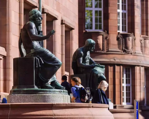 Vor einem roten Sandsteingebäude stehen bronzene Sitzstatuen, davor gehen mehrere Studierende vorbei.