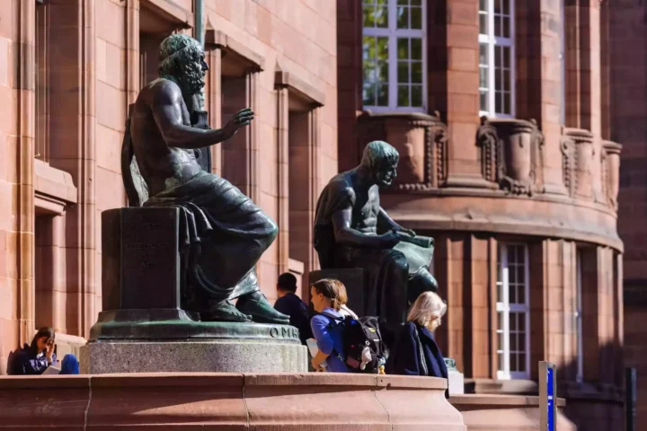 Vor einem roten Sandsteingebäude stehen bronzene Sitzstatuen, davor gehen mehrere Studierende vorbei.