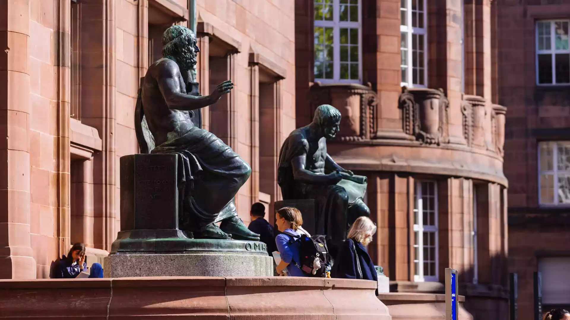 Vor einem roten Sandsteingebäude stehen bronzene Sitzstatuen, davor gehen mehrere Studierende vorbei.