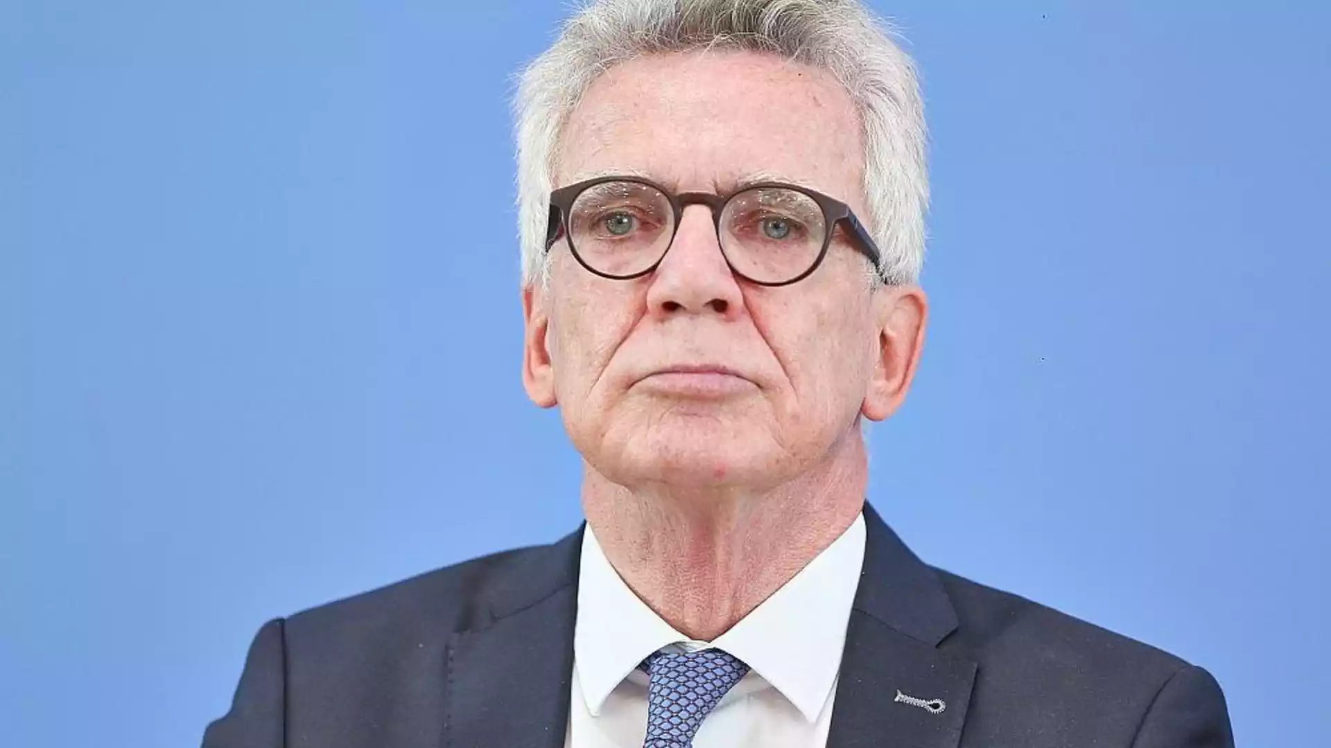 Porträt eines älteren Mannes mit Brille und grauen Haaren im Anzug vor blauem Hintergrund.