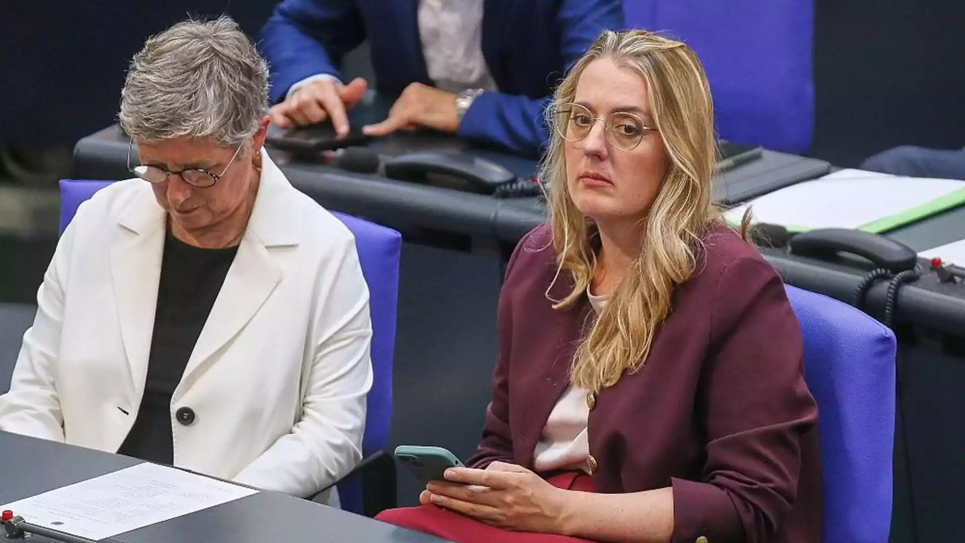 Zwei Politikerinnen sitzen in einer Sitzungsbank im Parlament, eine schaut auf ihr Smartphone.