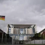 EU- und Deutschlandflagge vor einem Regierungsgebäude unter wolkigem Himmel.