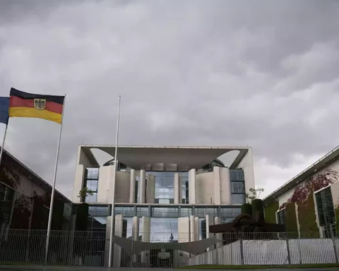 EU- und Deutschlandflagge vor einem Regierungsgebäude unter wolkigem Himmel.