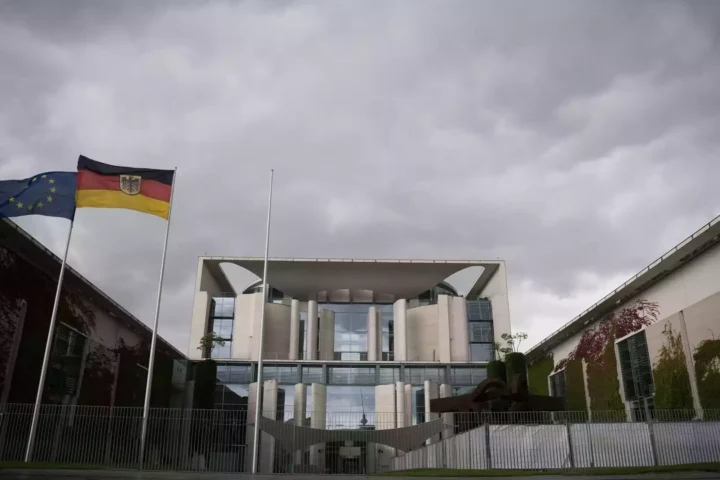 EU- und Deutschlandflagge vor einem Regierungsgebäude unter wolkigem Himmel.