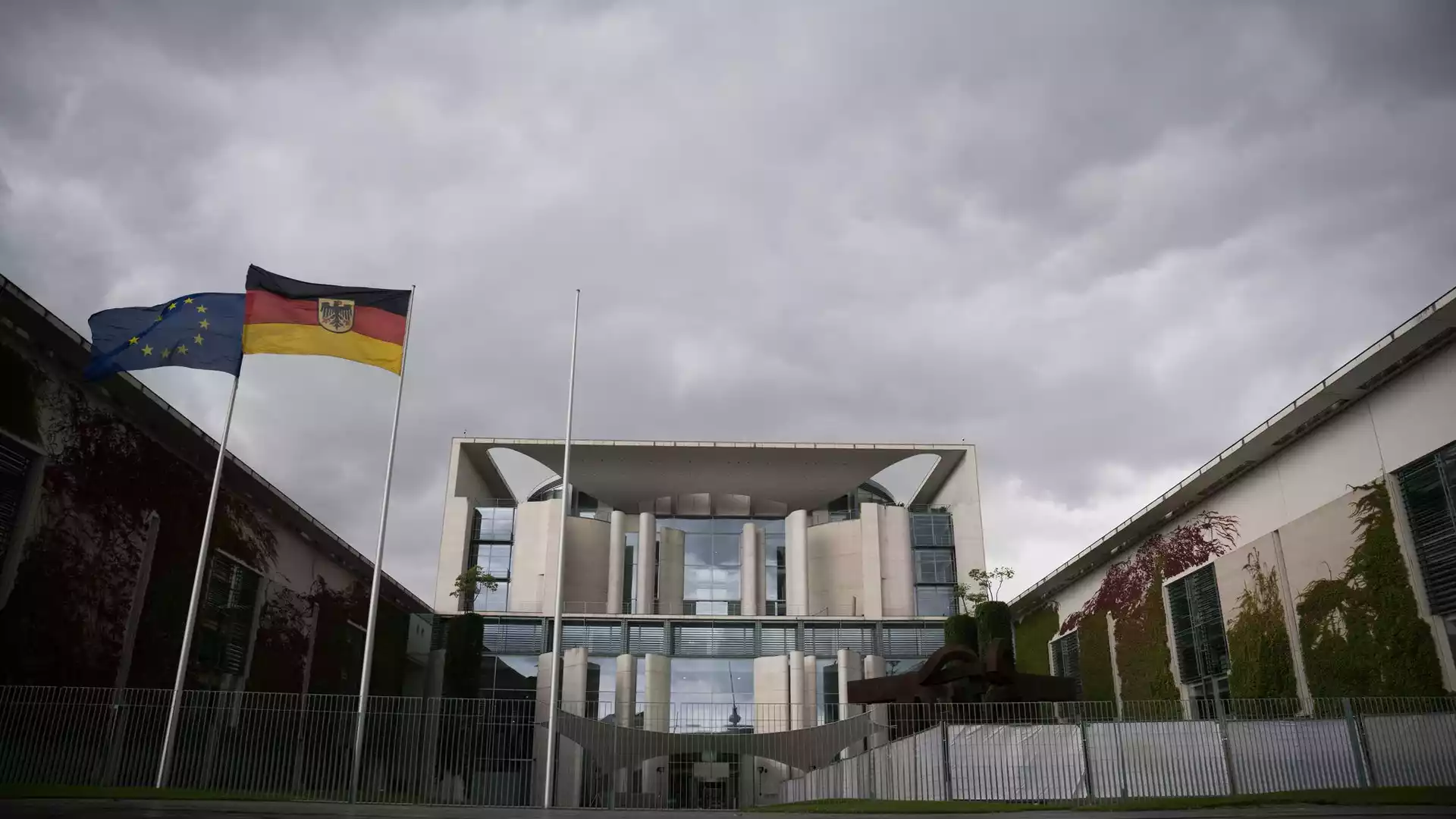 EU- und Deutschlandflagge vor einem Regierungsgebäude unter wolkigem Himmel.