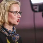 Eine blonde Frau mit Brille spricht in einer Studioaufnahme im Profil.