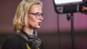 Eine blonde Frau mit Brille spricht in einer Studioaufnahme im Profil.