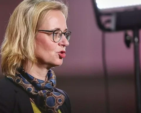 Eine blonde Frau mit Brille spricht in einer Studioaufnahme im Profil.