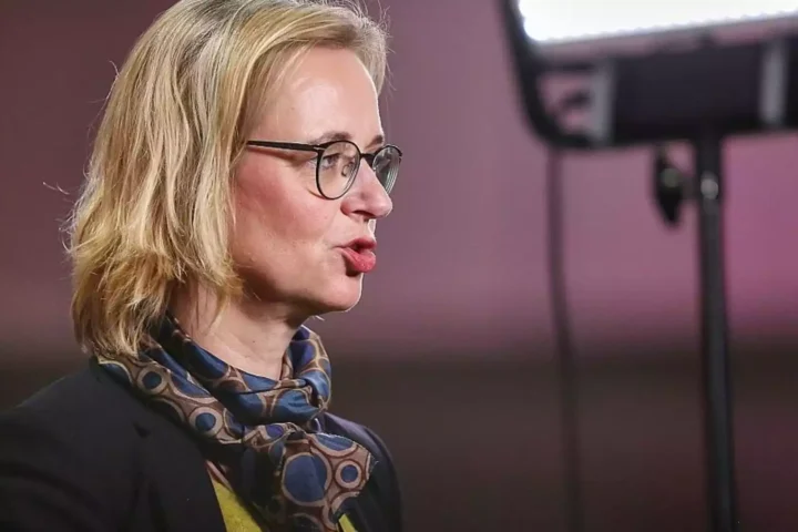 Eine blonde Frau mit Brille spricht in einer Studioaufnahme im Profil.