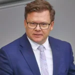 Ein Mann in blauem Anzug und Brille spricht vor grauem Hintergrund.