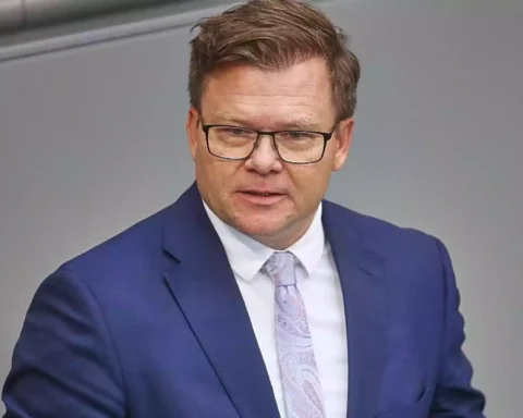 Ein Mann in blauem Anzug und Brille spricht vor grauem Hintergrund.