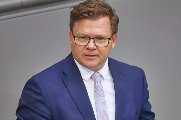 Ein Mann in blauem Anzug und Brille spricht vor grauem Hintergrund.