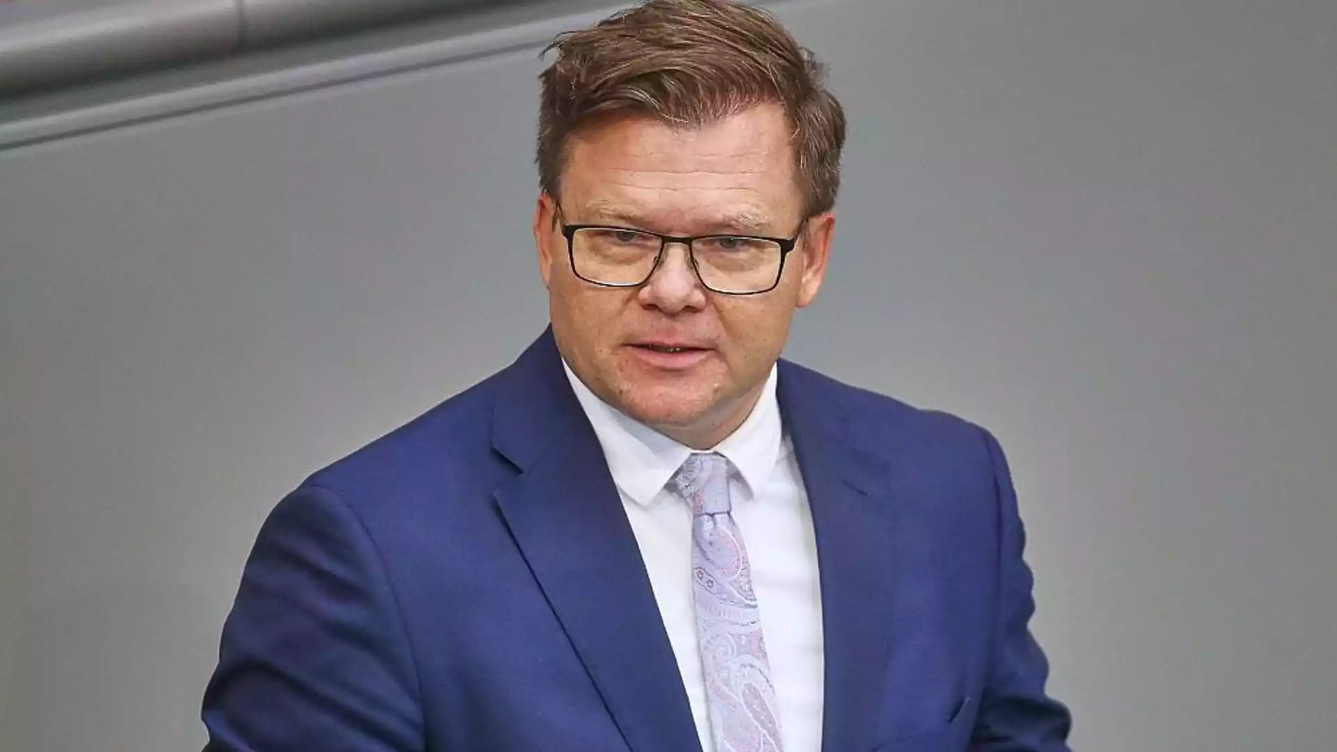Ein Mann in blauem Anzug und Brille spricht vor grauem Hintergrund.