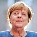 Angela Merkel im Porträt vor grauem Hintergrund mit hellem Gegenlicht.
