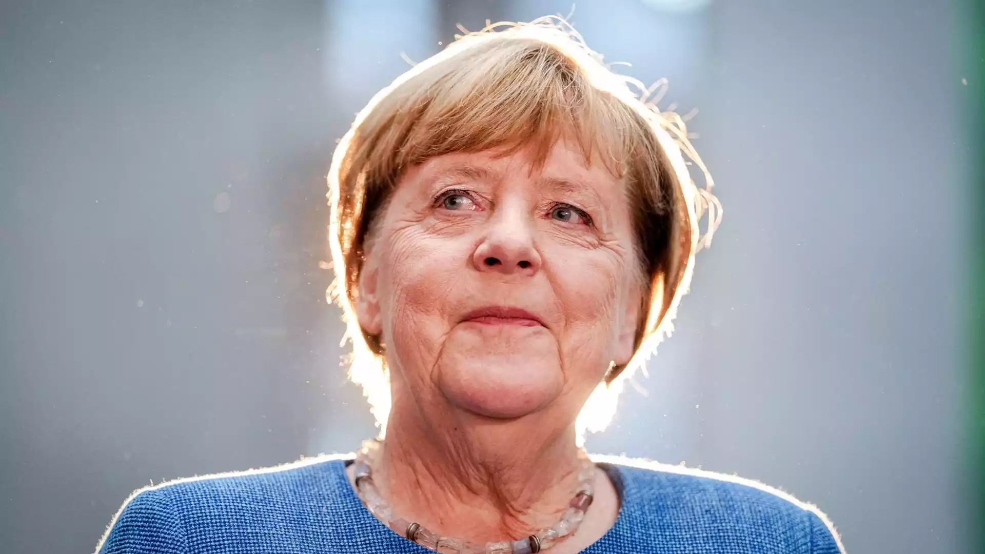 Angela Merkel im Porträt vor grauem Hintergrund mit hellem Gegenlicht.