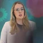 Eine Frau mit Brille spricht vor einem grünen Hintergrund mit verschwommenen Logos.