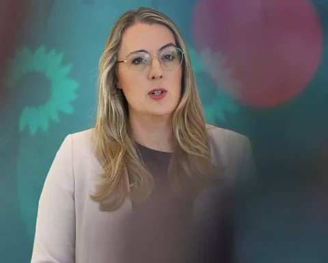 Eine Frau mit Brille spricht vor einem grünen Hintergrund mit verschwommenen Logos.