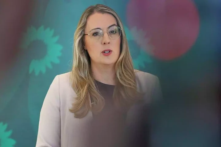 Eine Frau mit Brille spricht vor einem grünen Hintergrund mit verschwommenen Logos.