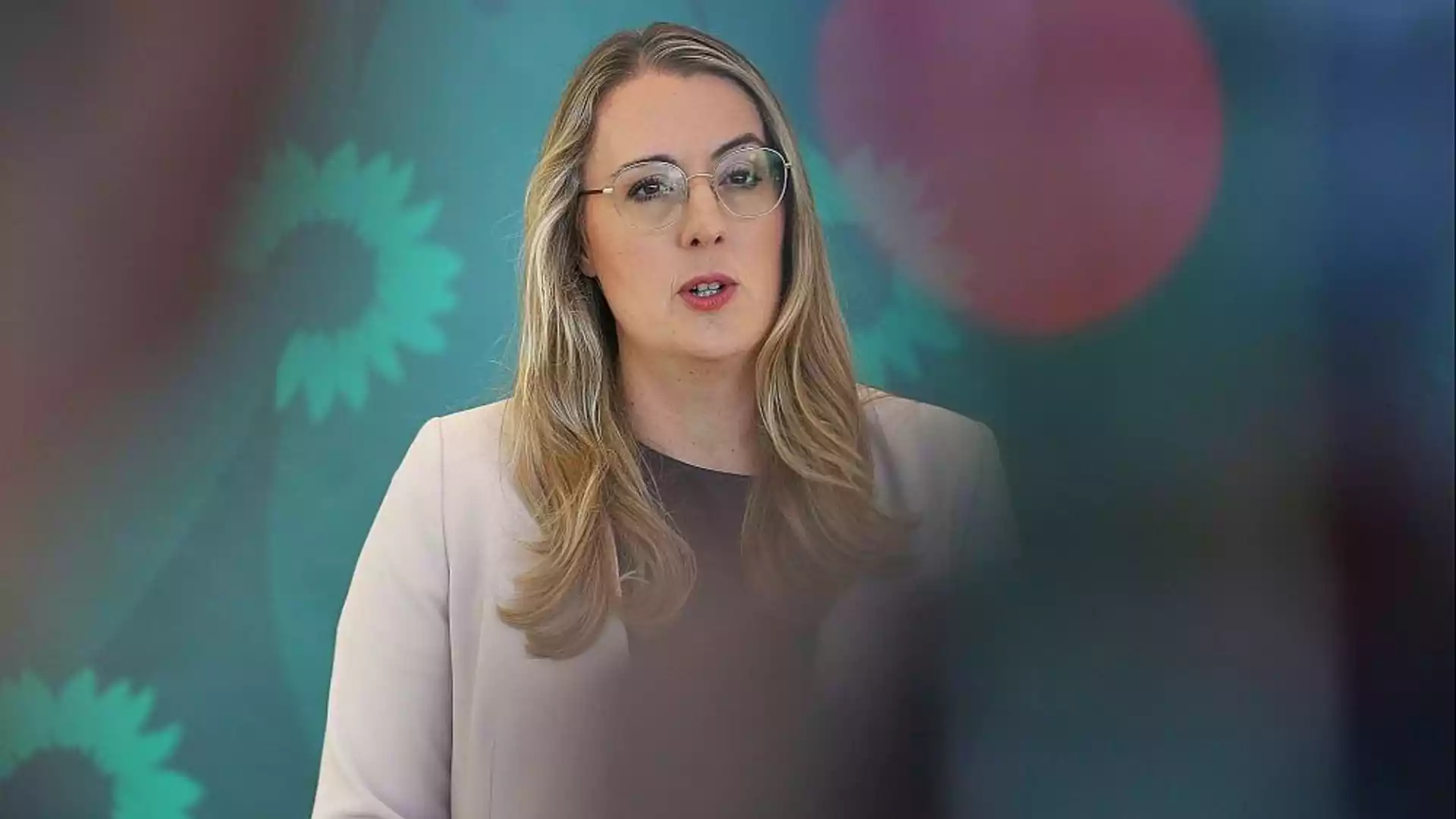 Eine Frau mit Brille spricht vor einem grünen Hintergrund mit verschwommenen Logos.