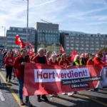 Demonstrierende mit rotem Banner und mehreren Fahnen auf einer Straße in einer Stadt.