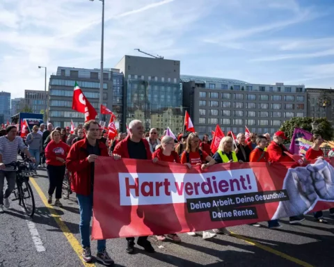 Demonstrierende mit rotem Banner und mehreren Fahnen auf einer Straße in einer Stadt.