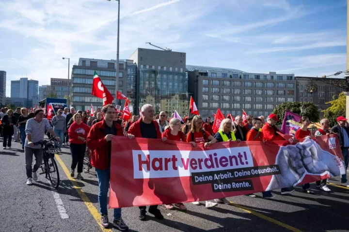 Demonstrierende mit rotem Banner und mehreren Fahnen auf einer Straße in einer Stadt.