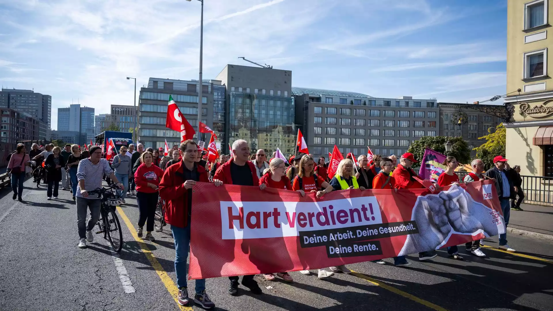 Bündnis aus Verbänden und Gewerkschaften warnt vor Kürzungen in der Pflege 1 Demonstrierende mit rotem Banner und mehreren Fahnen auf einer Straße in einer Stadt.