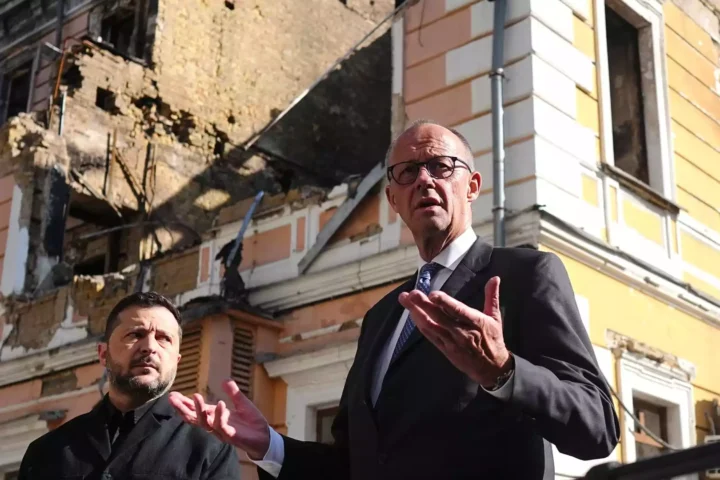 Friedrich Merz spricht vor einem beschädigten Gebäude in Kiew, neben ihm steht Wolodymyr Selenskyj.