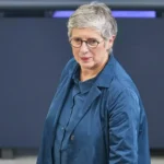 Eine Frau mit kurzen grauen Haaren und Brille steht in einem blauen Outfit vor einer dunklen Wand.